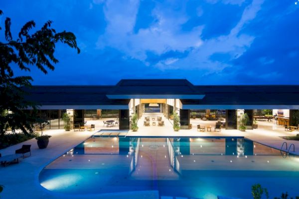 The image shows a luxurious modern building with an illuminated pool and open seating area under a twilight sky.