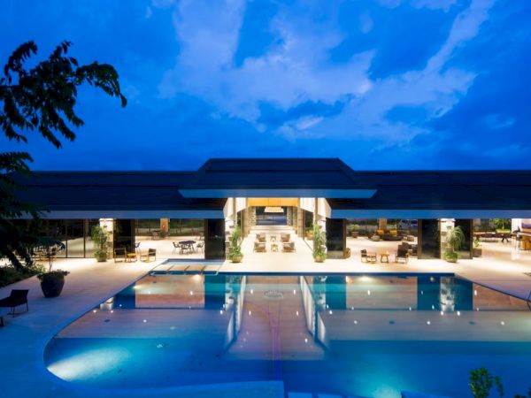 The image shows a luxurious modern building with an illuminated pool and open seating area under a twilight sky.