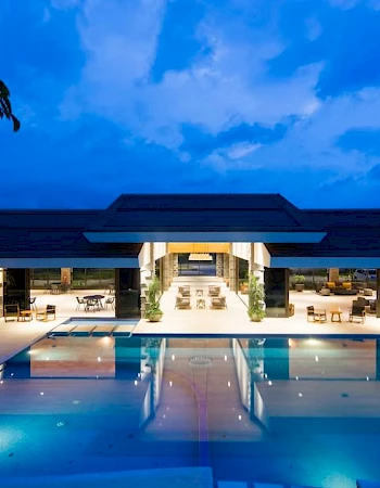 A modern building with a pool at dusk; interior lights create a warm glow.