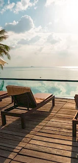 A serene poolside with lounge chairs, umbrellas, and palm trees, overlooking a calm sea under a partly cloudy sky.