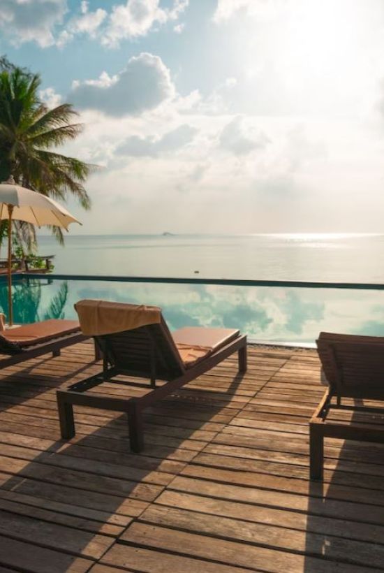 A wooden deck with lounge chairs and umbrellas overlooks an infinity pool, with palm trees and a calm sea in the background.
