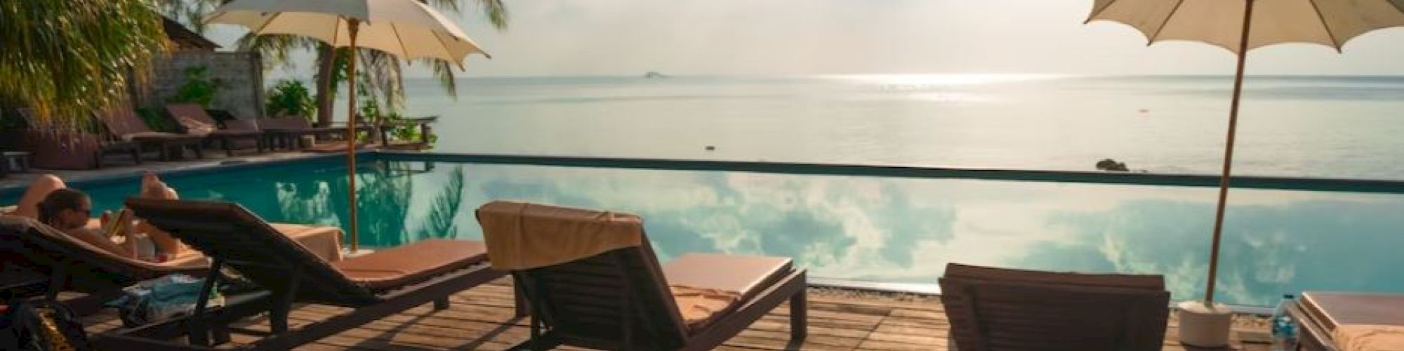 A tranquil poolside scene with lounge chairs, umbrellas, and palm trees overlooking a serene body of water under a partly cloudy sky.