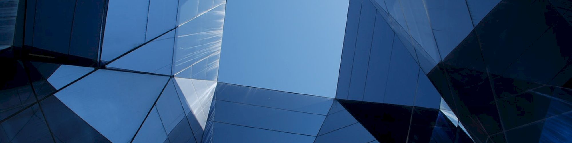 The image shows a geometric architectural structure with reflective glass surfaces converging towards an open sky.