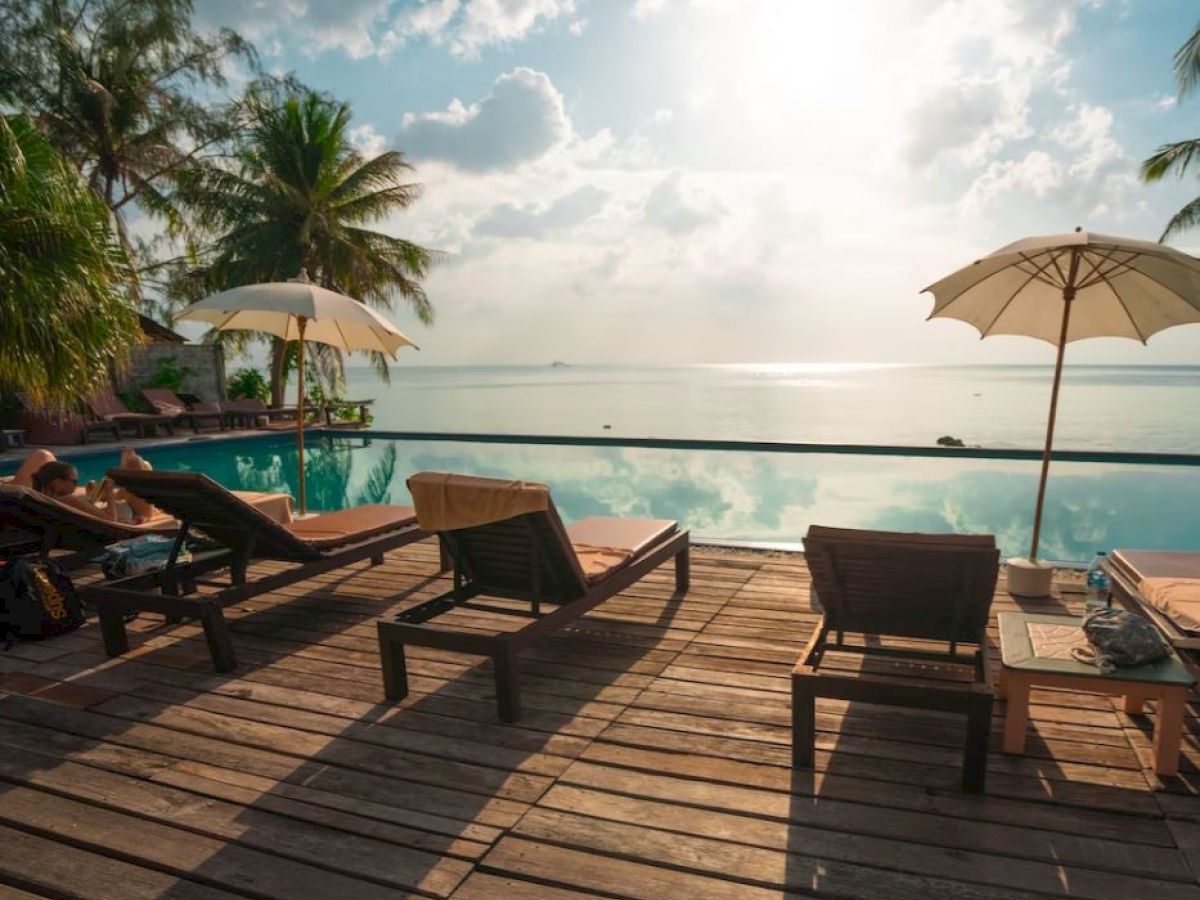 Poolside deck with lounge chairs, umbrellas, and palm trees overlooking a tranquil sea under a partly cloudy sky at sunset.