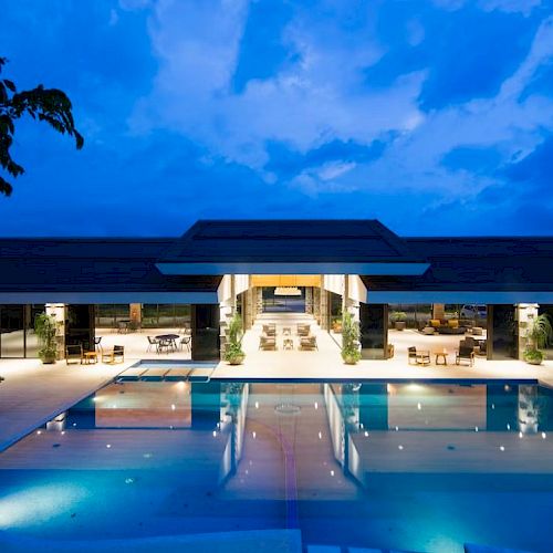 A luxurious modern building with a lit swimming pool, surrounded by outdoor seating, set against a deep blue evening sky.