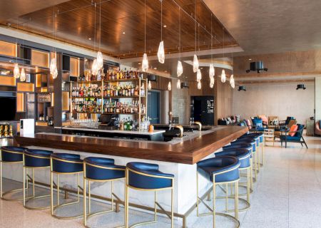 A stylish bar features a wooden countertop, pendant lights, blue chairs, and a well-stocked back bar, creating a modern lounge atmosphere.