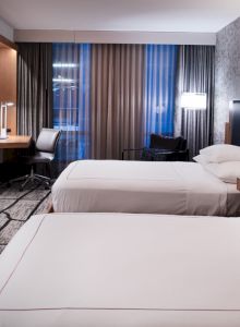 A modern hotel room with two beds, a wall-mounted TV, a desk, and patterned carpet. The room is well-lit with decorative lamps.