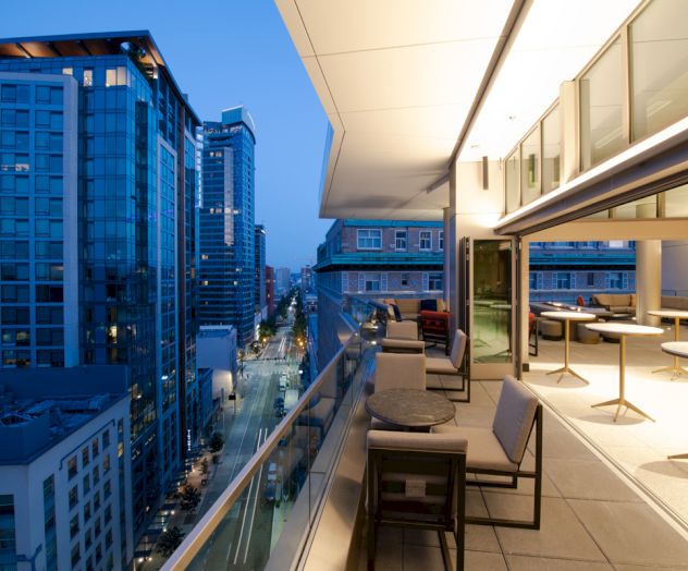 A modern balcony overlooks a cityscape with tall buildings at dusk, featuring outdoor seating and tables along the railing.