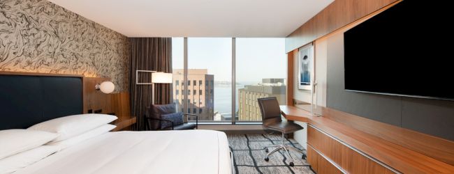 A modern hotel room with a large bed, wall-mounted TV, desk, windows with a city view, and stylish decor.
