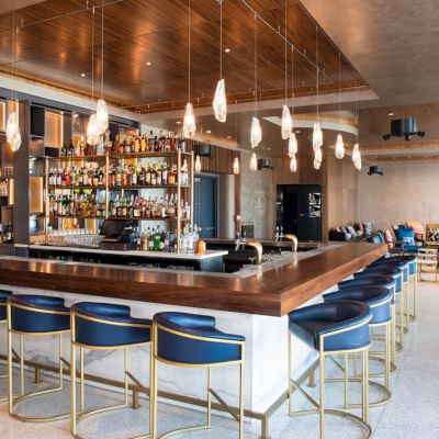 A modern bar with blue stools, a fully stocked bar, pendant lights, and a stylish lounge area in the background.