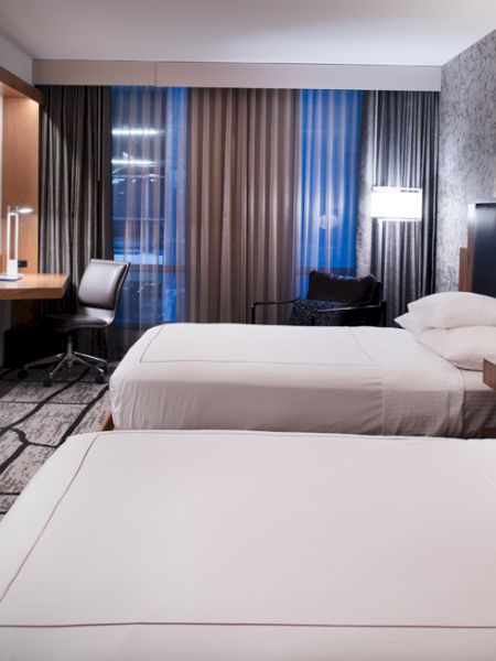 The image shows a modern hotel room with two beds, nightstands, a desk, chair, TV, and patterned wallpaper and carpet.