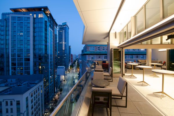 The image shows a modern cityscape at dusk, featuring high-rise buildings and a stylish balcony with seating and tables.