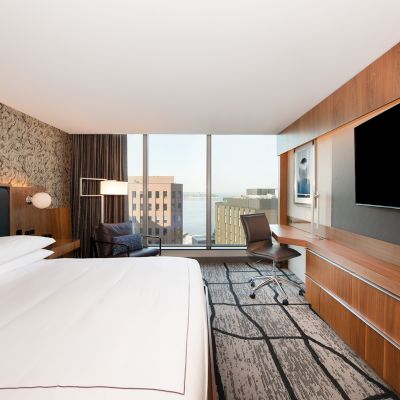 A modern hotel room with a large bed, wall-mounted TV, desk, chair, and a window offering a cityscape view.