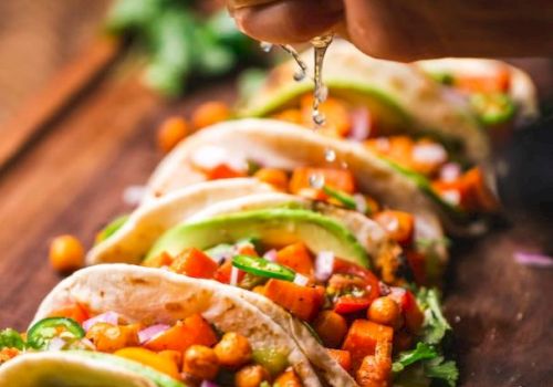 A hand squeezes lime juice over tacos filled with chickpeas, avocado, and vegetables on a wooden board. Lime slices are in the foreground.