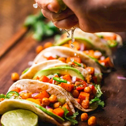 A hand squeezes lime juice over tacos filled with chickpeas, avocado, and vegetables on a wooden board. Lime slices are in the foreground.