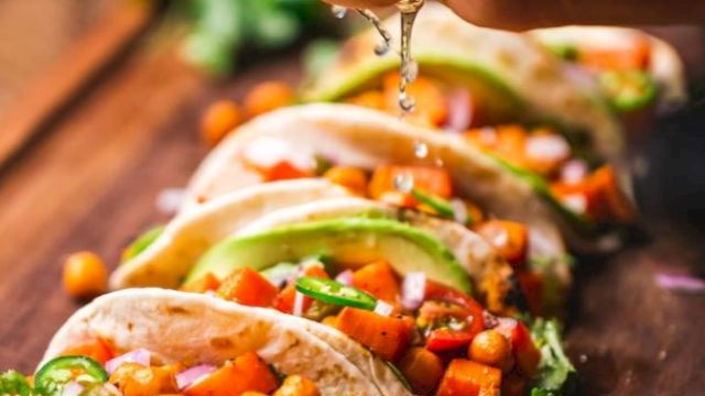 A hand squeezes lime juice over tacos filled with chickpeas, avocado, and vegetables on a wooden board. Lime slices are in the foreground.