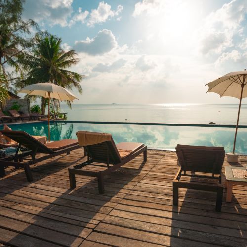 A serene poolside view with lounge chairs and umbrellas, overlooking a calm sea under a partly cloudy sky with sunlight shining through.