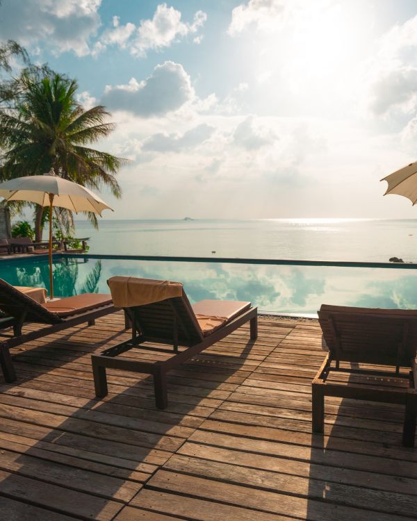 A serene poolside view with lounge chairs and umbrellas, overlooking a calm sea under a partly cloudy sky with sunlight shining through.