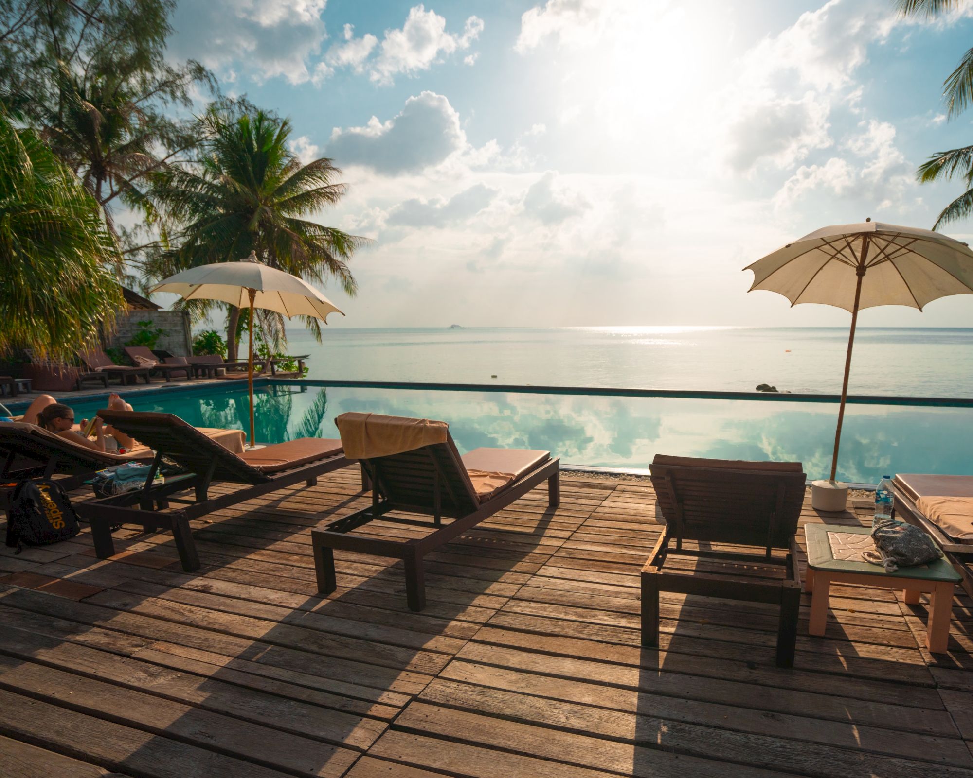 A serene poolside with lounge chairs, umbrellas, palm trees, and a view of the ocean under a bright sky.