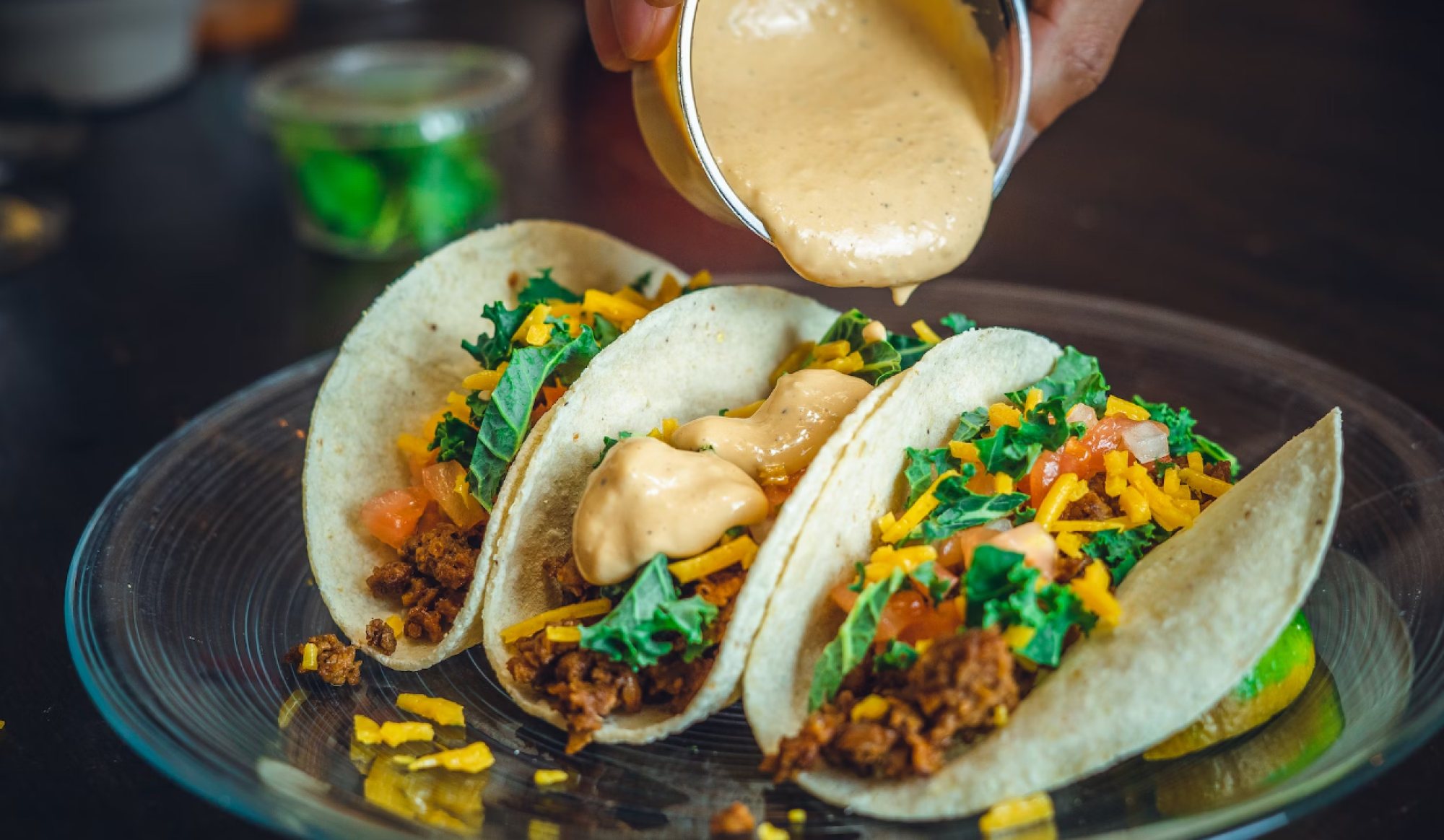 Three tacos with lettuce, cheese, and sauce being poured on them, placed on a glass plate.