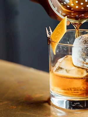 A cocktail being poured into a glass with ice and a lemon twist garnish on the rim.