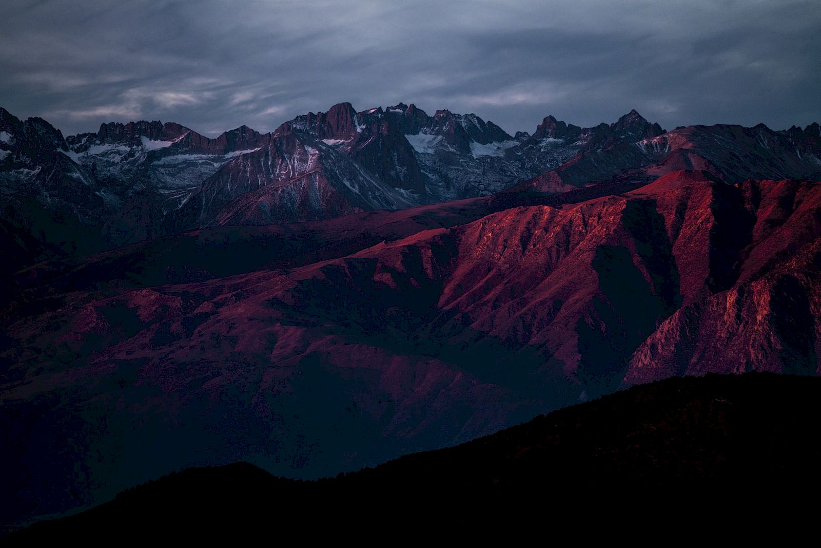 Mountains under a dramatic sky with peaks bathed in red-orange light during twilight, casting shadows on the landscape.