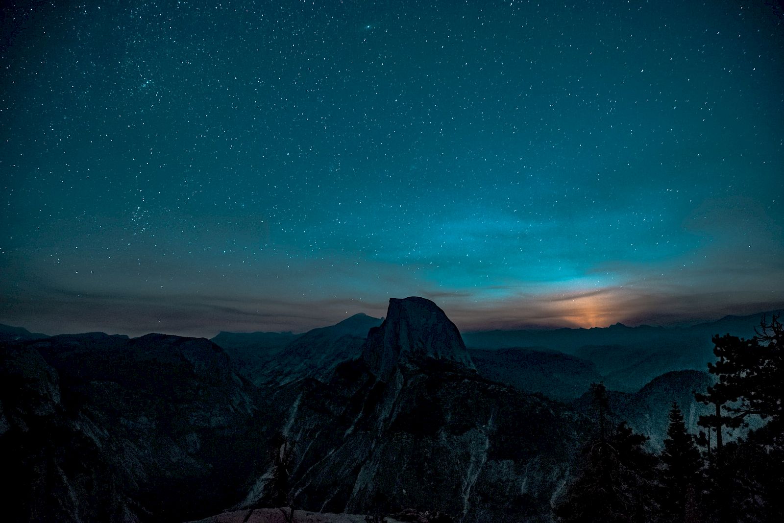 A starry night sky over a mountain landscape with a glowing horizon, possibly from a setting sun or moon, creating a serene scene.