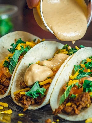 Three tacos with meat, cheese, lettuce, and sauce being poured over.