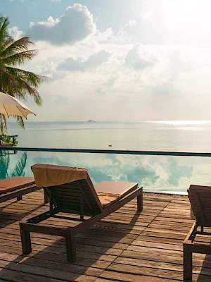 Sun loungers by an infinity pool overlooking the ocean, surrounded by palm trees and umbrellas, with a serene, sunny atmosphere.