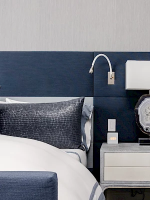 A modern bedroom with a navy and gray color scheme, featuring a bed, pillows, a nightstand, and a lamp on a decorative stone base.