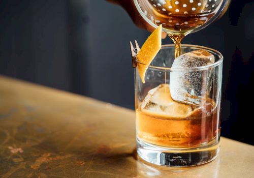 A hand pours a brown liquid into a glass with ice and a lemon twist garnish, likely a cocktail, on a wooden table.
