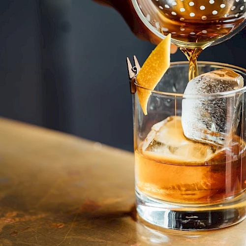 A hand pours a brown liquid into a glass with ice and a lemon twist garnish, likely a cocktail, on a wooden table.