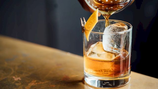 A hand pours a brown liquid into a glass with ice and a lemon twist garnish, likely a cocktail, on a wooden table.