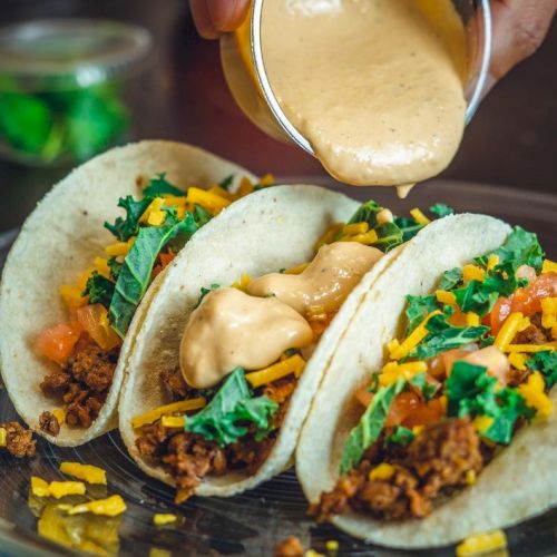 Three tacos filled with ground meat, cheese, lettuce, and tomato, being topped with sauce from a cup.
