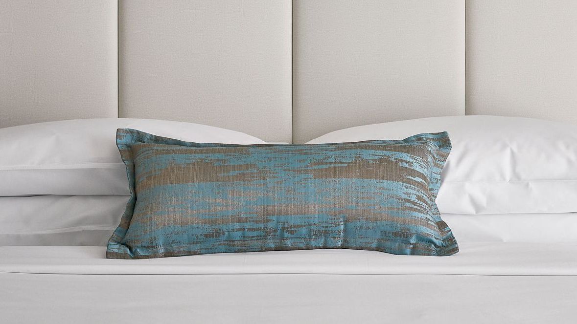 A neatly made bed with white pillows and a blue and brown accent pillow is shown against a light-colored headboard.