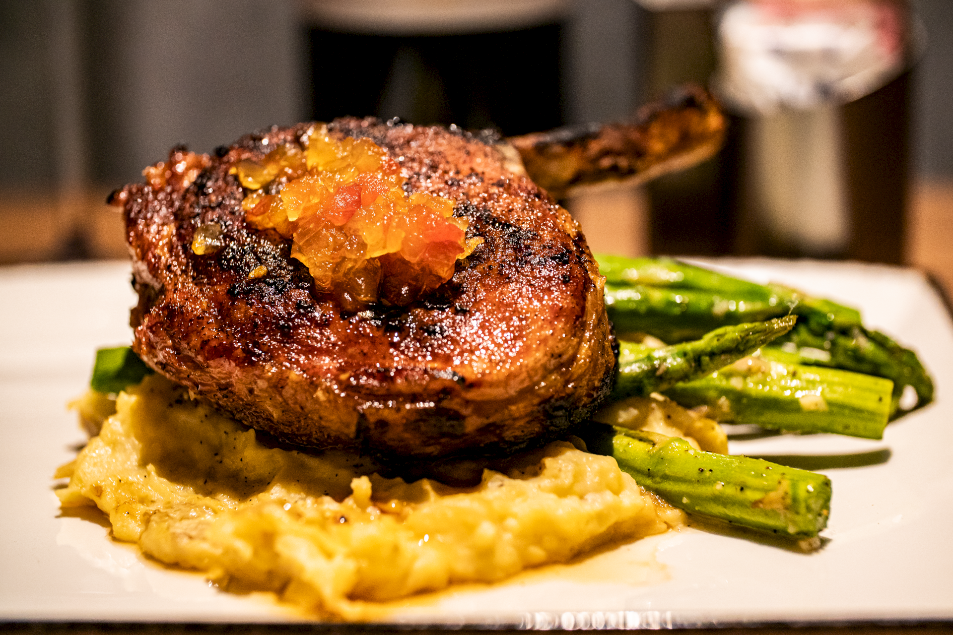 A grilled pork chop with sauce on top, served with mashed potatoes and asparagus on a white plate.