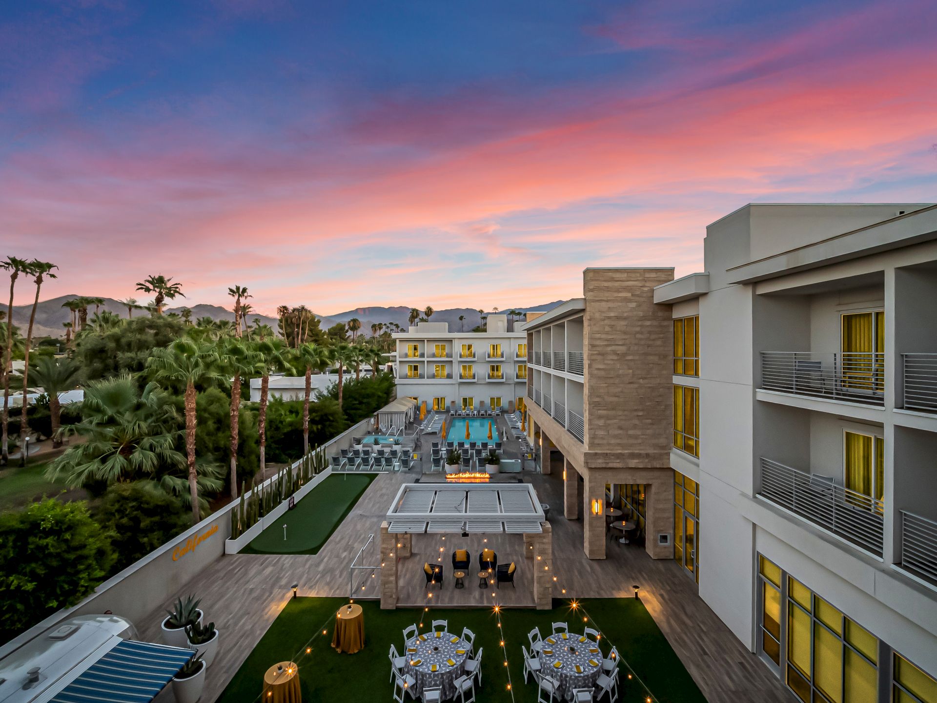A luxury hotel courtyard with a pool, lounge chairs, and festive lights at sunset, surrounded by palm trees and mountains.