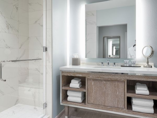 The image shows a modern bathroom with a shower, a large mirror, a sink, and shelves stocked with white towels underneath the vanity counter.