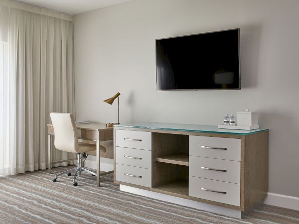 The image shows a modern hotel room with a desk, chair, lamp, TV, and dresser. The decor is minimalistic and neutral in tone.