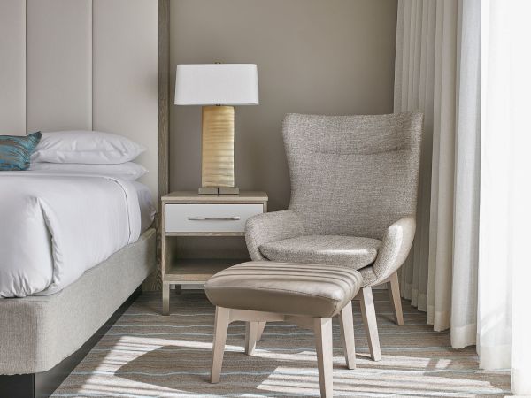 A cozy room with a bed, a nightstand with a lamp, and an armchair with an ottoman, all in neutral tones by a window.