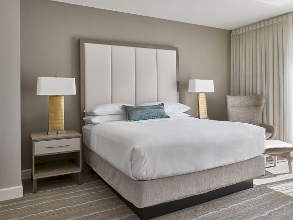 A modern bedroom with a queen bed, two bedside tables with lamps, a chair, and curtains, all in neutral tones, creating a serene atmosphere.