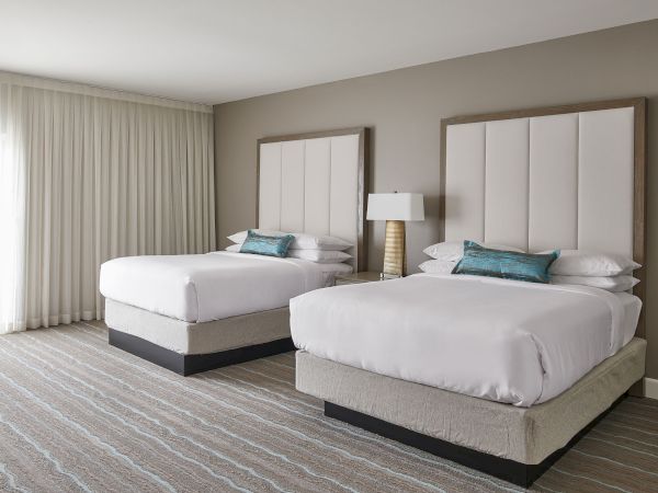 The image shows a hotel room with two neatly made beds, a lamp between them, striped carpet, and curtains letting in soft light.