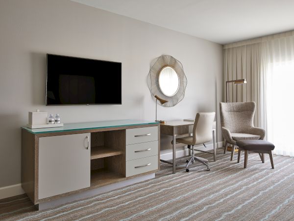 A modern hotel room interior with a TV, desk, chair, wall mirror, and armchair. Bright and minimalistic design with neutral tones.