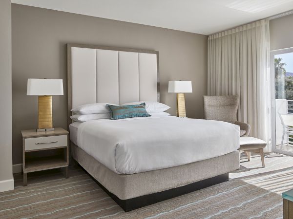 The image shows a modern hotel room with a large bed, two bedside tables with lamps, a chair, and sheer curtains covering a balcony door.