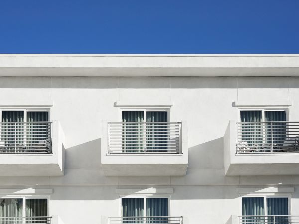 The image shows a minimalist building facade with three identical balconies featuring railing and glass doors against a clear blue sky.
