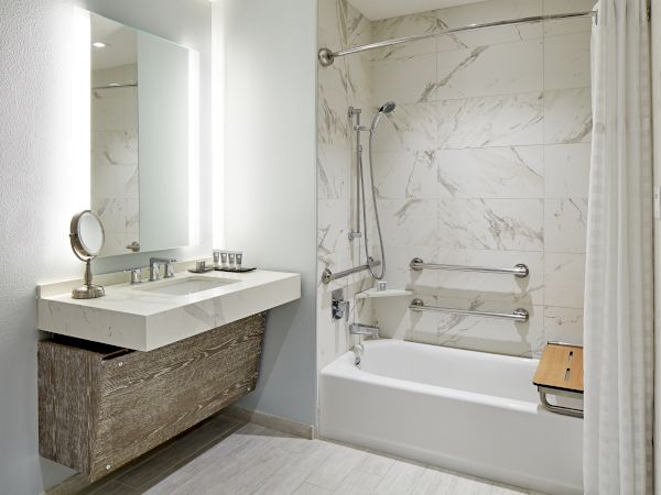 A bathroom with a countertop, sink, mirror, tiled wall, bathtub, grab bars, and a shower curtain in neutral tones.