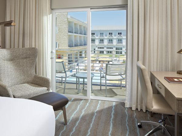 The image shows a modern hotel room with a bed, armchair, desk, and a balcony with outdoor seating overlooking other hotel buildings.
