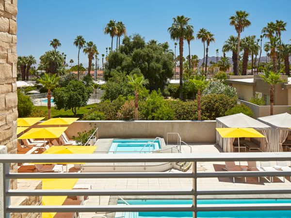 A sunny outdoor pool area with yellow umbrellas, loungers, and cabanas, surrounded by trees and palm trees.
