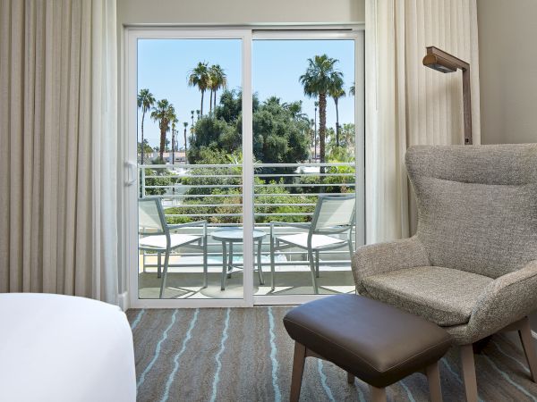 A cozy room with an armchair and ottoman faces a balcony with chairs, overlooking palm trees through sliding glass doors.