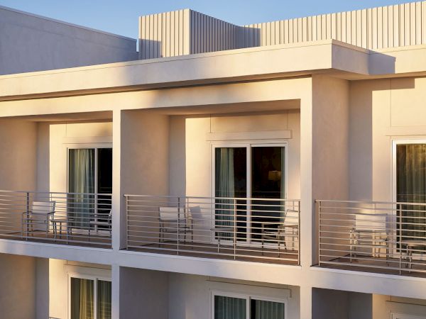 The image shows a modern building with balconies and sliding glass doors, featuring metal railings and outdoor seating.